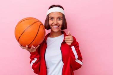 Genç melez kadın basketbol oynuyor. Pembe arka planda soyutlanmış. Gülümsüyor ve başparmağını kaldırıyor.