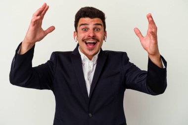 Beyaz arka planda izole edilmiş kablosuz kulaklık takan genç beyaz iş adamı hoş bir sürpriz alıyor, heyecanlı ve el kaldırıyor..