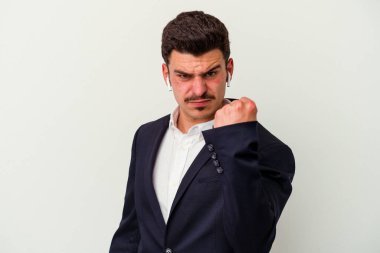 Beyaz arka planda izole edilmiş kablosuz kulaklık takan genç beyaz iş adamı kameraya yumruk gösteriyor, agresif yüz ifadesi.