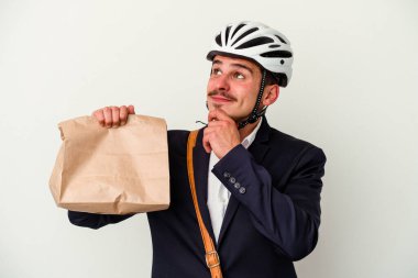 Bisiklet kaskı takan ve elinde tuttuğu genç beyaz arka planda izole edilmiş yiyecekleri kuşkulu ve şüpheci bir ifadeyle yana bakan beyaz iş adamı..