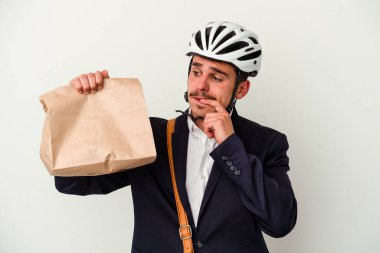 Bisiklet kaskı takan ve elinde tuttuğu beyaz arka planda izole edilmiş yiyecekleri alan beyaz iş adamı rahat bir şekilde fotokopi odasına bakan bir şeyi düşünüyor..