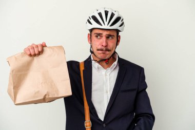 Bisiklet kaskı takan ve elinde tuttuğu beyaz arka planda izole edilmiş yiyecekleri alan genç beyaz bir adam kafası karışmış, şüpheli ve güvensiz hissediyor..