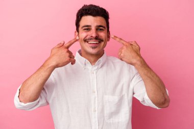 Pembe arka planda izole edilmiş genç beyaz adam gülümsüyor, parmaklarını ağıza doğrultuyor..