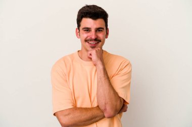 Beyaz arka planda izole olmuş genç beyaz bir adam gülümsüyor ve kendinden emin, çenesine elleriyle dokunuyor..