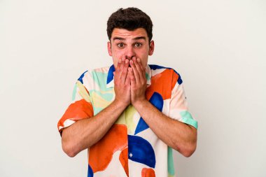 Beyaz arka planda izole edilmiş genç beyaz adam elleriyle ağzını kapatırken şok oldu..