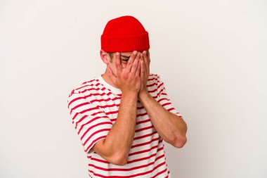 Beyaz arka planda izole edilmiş genç beyaz bir adam parmaklarının arasından korkmuş ve gergin bir şekilde göz kırpıyor..