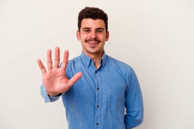 Beyaz arka planda izole edilmiş genç beyaz bir adam gülümseyerek beş numarayı parmaklarıyla gösteriyor..