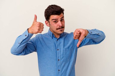 Beyaz arka planda izole edilmiş genç beyaz bir adam baş parmaklarını aşağı ve yukarı kaldırıyor, zor seçim konsepti