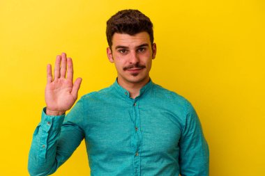 Sarı arka planda izole edilmiş genç beyaz adam gülümseyerek beş numarayı parmaklarıyla gösteriyor..
