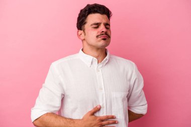 Pembe arka planda izole edilmiş genç beyaz adam karnına dokunuyor, nazikçe gülümsüyor, yemek yiyor ve tatmin oluyor..