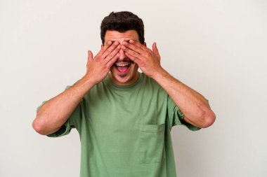 Beyaz arka planda soyutlanmış beyaz tenli genç bir adam elleriyle gözlerini kapatıyor, geniş bir gülümsemeyle bir sürpriz bekliyor..