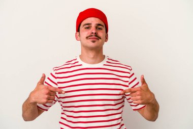 Beyaz arka planda yaşayan beyaz tenli genç bir adam gömlek fotokopi alanını gösteriyor, gururlu ve kendinden emin.