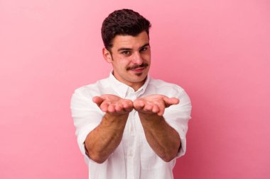 Pembe arka planda izole edilmiş genç beyaz bir adam elinde avuç içi bir şeyle kameraya sunuyor..