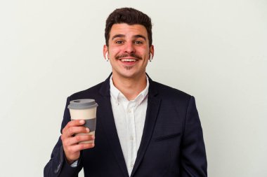 Kablosuz kulaklık takan ve elinde tuttuğu beyaz arka planda yalnız kahve içen mutlu, gülümseyen ve neşeli genç beyaz iş adamı..