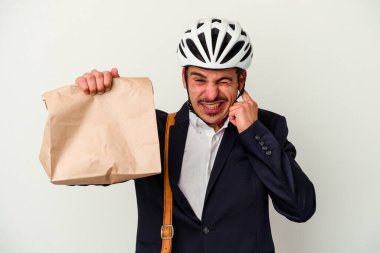 Bisiklet kaskı takan ve elinde tuttuğu beyaz arka planda izole edilmiş yiyecekleri alıp kulaklarını elleriyle kapatan genç beyaz iş adamı..