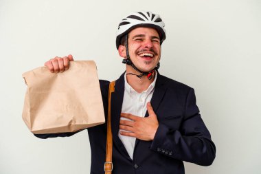 Bisiklet kaskı takan ve elinde beyaz arka planda izole edilmiş yiyecekler tutan beyaz iş adamı kahkahalarla gülüyor ve elini göğsüne koyuyor..