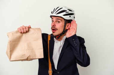 Bisiklet kaskı takan ve elinde tuttuğu beyaz atlı genç bir adam bir dedikoduyu dinlemek için beyaz arka planda izole edilmiş yiyecekleri alıyor..