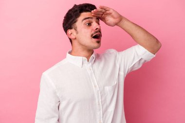Pembe arka planda izole edilmiş genç beyaz adam uzaklara bakıyor ve elini alnına koyuyor..