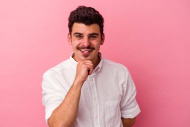 Pembe arka planda izole edilmiş genç beyaz adam gülümsüyor ve kendinden emin, elleriyle çenesine dokunuyor..