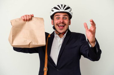 Bisiklet kaskı takan ve elinde tuttuğu beyaz arka planda izole edilmiş yiyecekleri alan genç beyaz bir adam hoş bir sürpriz yaparak heyecanlı ve el kaldırıyor..