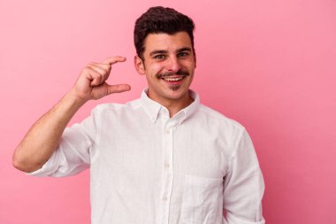 Pembe arka planda izole edilmiş genç beyaz adam işaret parmaklarıyla küçük bir şey tutuyor, gülümsüyor ve kendinden emin..
