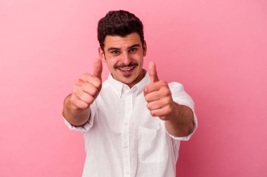 Pembe arka planda soyutlanmış genç beyaz bir adam. Başparmakları havada, bir şey için tezahürat yapıyor. Destek ve saygı kavramı..