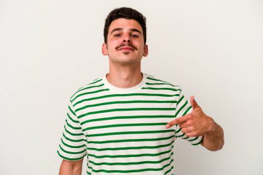 Beyaz arka planda yaşayan beyaz tenli genç bir adam gömlek fotokopi alanını gösteriyor, gururlu ve kendinden emin.