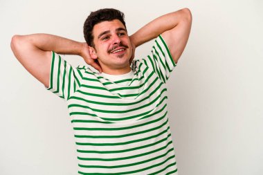 Beyaz arka planda izole olmuş genç beyaz bir adam kendini güvende hissediyor, elleri başının arkasında..