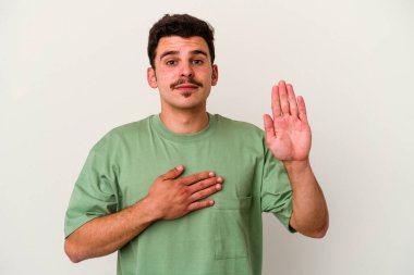 Beyaz arka planda izole edilmiş genç beyaz adam yemin ediyor, elini göğsüne koyuyor..