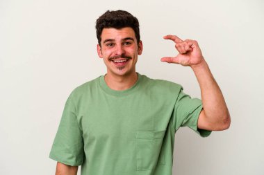 Beyaz arka planda izole edilmiş genç beyaz bir adam işaret parmaklarıyla küçük bir şey tutuyor, gülümsüyor ve kendinden emin..