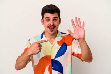 Beyaz arka planda izole edilmiş genç beyaz bir adam gülümseyerek beş numarayı parmaklarıyla gösteriyor..