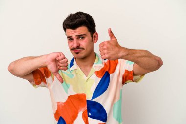 Beyaz arka planda izole edilmiş genç beyaz bir adam baş parmaklarını aşağı ve yukarı kaldırıyor, zor seçim konsepti