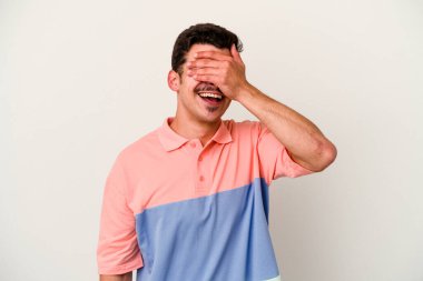 Beyaz arka planda soyutlanmış beyaz tenli genç bir adam elleriyle gözlerini kapatıyor, geniş bir gülümsemeyle bir sürpriz bekliyor..