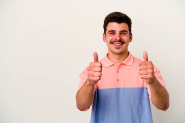 Beyaz arka planda izole edilmiş genç beyaz adam gülümsüyor ve başparmağını kaldırıyor.