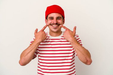 Beyaz arka planda izole edilmiş genç beyaz adam gülümsüyor, parmaklarını ağıza doğrultuyor..