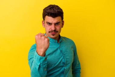 Sarı arka planda izole edilmiş genç beyaz adam kameraya yumruk gösteriyor, agresif yüz ifadesi var..