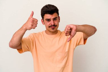 Beyaz arka planda izole edilmiş genç beyaz bir adam baş parmaklarını aşağı ve yukarı kaldırıyor, zor seçim konsepti