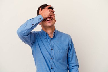 Beyaz arka planda soyutlanmış beyaz tenli genç bir adam elleriyle gözlerini kapatıyor, geniş bir gülümsemeyle bir sürpriz bekliyor..