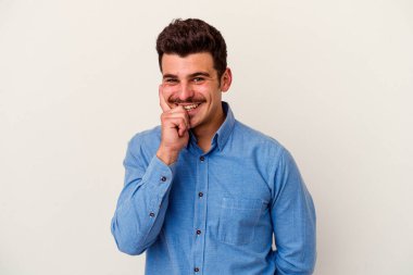 Beyaz arka planda izole olmuş genç beyaz bir adam gülümsüyor ve kendinden emin, çenesine elleriyle dokunuyor..