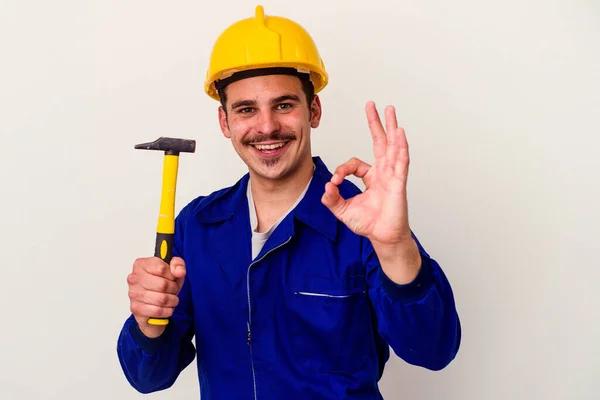 年轻的高加索工人拿着一把被白色背景隔离的铁锤快乐而自信地做出了一个好的手势 — 图库照片