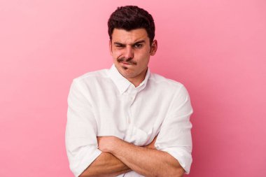 Pembe arka planda izole edilmiş genç beyaz adam hoşnutsuzca kaşlarını çatmış, kollarını kavuşturmuş..