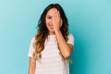 Mavi arka planda izole edilmiş genç Meksikalı kadın yüzünün yarısını avuç içiyle kaplayarak eğleniyor..