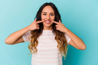 Mavi arka planda izole edilmiş genç Meksikalı kadın gülümsüyor, parmaklarını ağzına doğrultuyor..