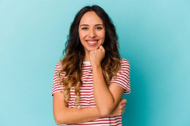 Mavi arka planda izole edilmiş genç Meksikalı kadın mutlu ve kendinden emin gülümsüyor, elleriyle çenesine dokunuyor..