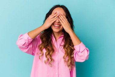 Mavi arka planda izole edilmiş genç Meksikalı kadın elleriyle gözlerini kapatıyor, gülümseyerek bir sürpriz bekliyor..