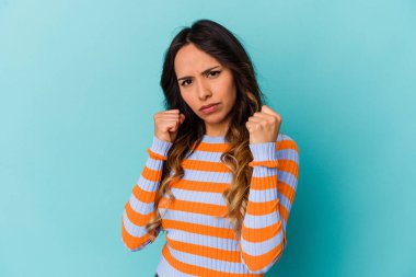 Mavi arka planda izole edilmiş genç Meksikalı kadın kameraya yumruk gösteriyor, agresif yüz ifadesi..