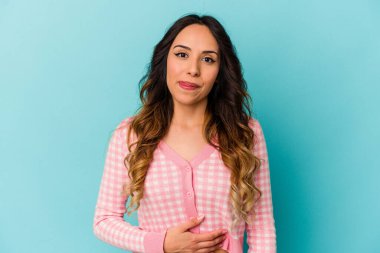 Mavi arka planda izole edilmiş genç Meksikalı kadın karnına dokunuyor, nazikçe gülümsüyor, yemek yiyor ve tatmin oluyor..