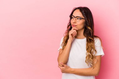 Pembe arka planda izole edilmiş genç Meksikalı kadın şüpheli ve şüpheci bir ifadeyle yana bakıyor..