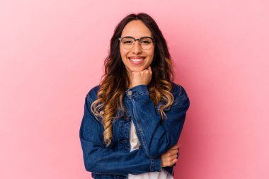 Pembe arka planda izole edilmiş genç Meksikalı kadın mutlu ve kendinden emin gülümsüyor, elleriyle çenesine dokunuyor..