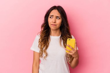 Pembe arka planda izole edilmiş cep telefonu tutan genç Meksikalı kadın şaşkın ve kararsız hissediyor..